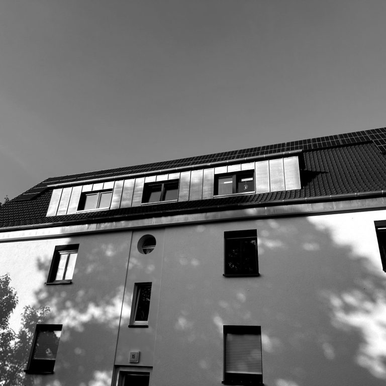 Dachfläche und Fassade eines Hauses in Paderborn. Das Bild zeigt eine helle Hausfassade mit Fenstern und Schattenwurf von Bäumen.
