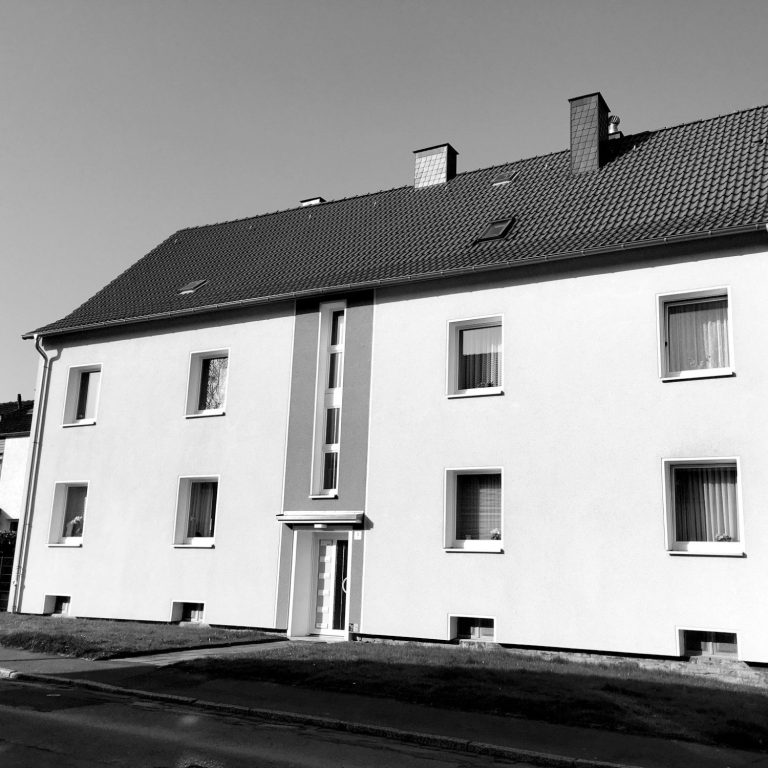 Hausfassade eines Vier-Familienhauses. Zweistöckiges, weißes Gebäude mit Fenster und einem zentralen Eingang.