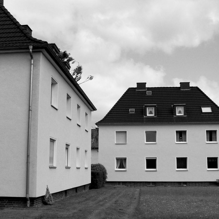 Garten und Fassaden einer Wohnanlage. Zwei weiße Gebäude mit Dachfenstern und Rasenfläche dazwischen.